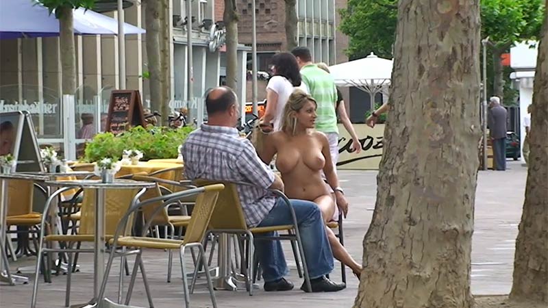 Brave, busty and mature naked woman sits brazenly at a crowded outdoor cafe